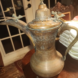 Puede incluir: Una cafetera de cobre antigua con grabados intrincados y remates decorativos en forma de pájaro. La cafetera tiene un cuerpo redondeado y un pico alto.