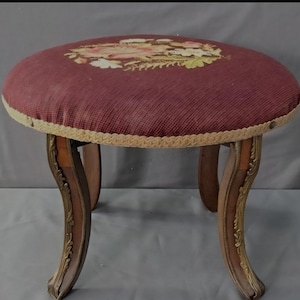 Puede incluir: Un taburete redondo antiguo con un asiento de tela burdeos con un diseño floral. El asiento está ribeteado con una cuerda de color crema. Las patas curvas son de madera con adornos metálicos dorados.