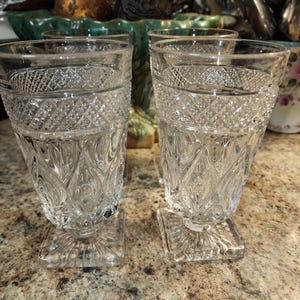 May include: Four clear glass tumblers with a cut glass design and square bases. The glasses are empty and sitting on a brown countertop.
