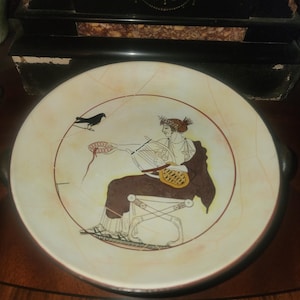 May include: A round, cream-colored ceramic plate with a hand-painted classical scene. The design features a seated figure playing a stringed instrument, a bird, and a red border. The plate has two black handles.