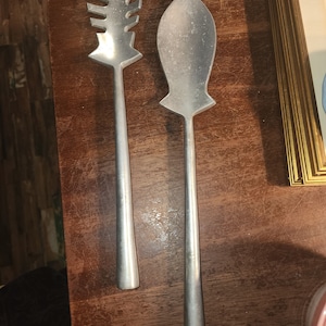 Puede incluir: Dos utensilios de servir de plata con forma de pez. El utensilio de la izquierda tiene un borde dentado y el utensilio de la derecha es liso.