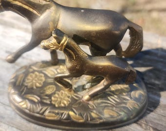 May include: A bronze-colored metal sculpture of a mare and foal. The mare stands behind the foal, which is in mid-stride. The sculpture is mounted on a round base with floral detailing.