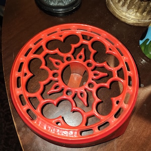 Puede incluir: Un salvamanteles redondo de hierro fundido de color naranja brillante con un intrincado diseño de celosía calada y un motivo central en forma de estrella. La palabra LE CREUSET está grabada en el borde exterior del marco circular.