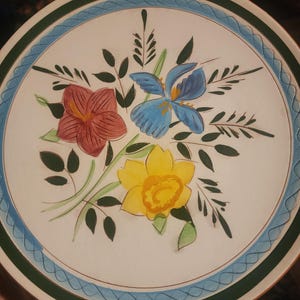 Puede incluir: Plato redondo de cerámica con diseño floral. El plato presenta un fondo blanco con un ramo pintado de flores rojas, azules y amarillas. Un borde verde y azul enmarca el diseño floral.