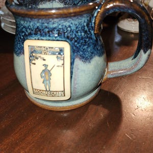 May include: A handcrafted ceramic mug with a blue and brown glaze. The mug features a rectangular emblem with an illustration of a person and the text "Moores Creek National Battlefield". The handle is curved and the mug sits on a small base.