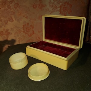 May include: A cream-coloured rectangular box with a red velvet interior, open to reveal its contents. Two small, cream-coloured, round containers sit beside the box. The items are on a dark green surface.