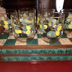 May include: Six clear drinking glasses, each decorated with a lemon and leaf motif. The glasses sit on a wooden tray with a green and white chequered pattern. The tray has a decorative green trim.