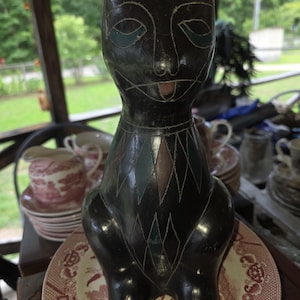 May include: A dark brown, carved wooden cat figurine featuring etched, diamond-shaped patterns on its chest and neck, with inlaid teal and reddish-brown accents on the eyes, nose, and chest, standing upright with a smooth, polished finish.
