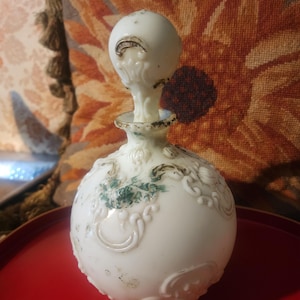 May include: A white, antique perfume bottle with a round body and a decorative stopper. The bottle features ornate embossed detailing and accents of green and gold. It sits on a red tray.
