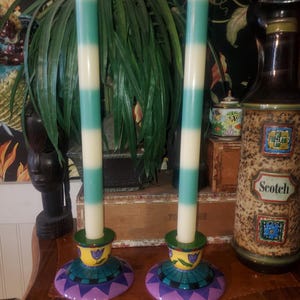 May include: Two tall, striped candles in teal and white, set in decorative candle holders. The holders feature a yellow, teal, and purple design. A bottle of Scotch is visible in the background.