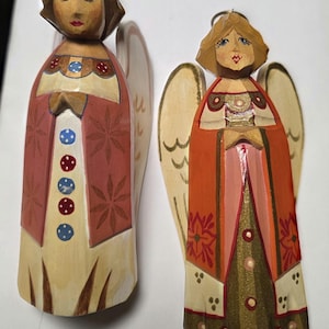 May include: Two hand-carved wooden angel figurines with painted details. The left angel wears a cream robe with red panels, blue and red dots, and star patterns. The right angel features an orange robe with gold accents, floral motifs, and a metallic base.
