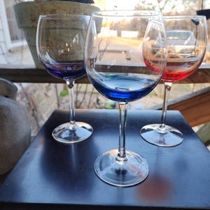May include: Three stemmed wine glasses with colored bases in purple, blue, and red. The glasses are on a black surface, with a blurred outdoor background. A turtle statue sits in a pot in the background.