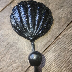 May include: A black seashell-shaped wall hook. The hook is made of a glossy, dark material and features a detailed shell design. The hook has a rounded knob and a stem for mounting. Decorative and functional.