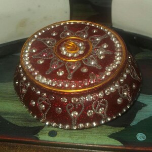 May include: A round, red jewelry box with a gold rim and intricate silver and clear crystal embellishments. The lid features a sunburst design with a central gold spiral. The box is on a green surface.