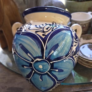 Könnte beinhalten: Eine Keramikvase mit cremefarbenem Korpus und blauem Blumenmuster. Die Vase hat zwei kleine Henkel und einen dunkelblauen Rand. Das Blumenmuster weist hellblaue und dunkelblaue Pinselstriche auf.