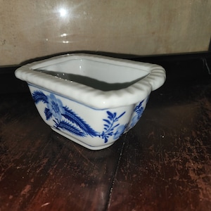 May include: A small, white ceramic planter with a blue and white floral design. The planter has a rectangular shape with rounded edges and a slightly raised rim.