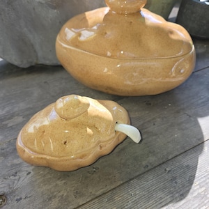 May include: Two-piece ceramic serving dish set. The set includes a lidded serving bowl and a smaller dish with a spoon. Both pieces are a speckled tan color with a glossy finish. The lid of the bowl has a small knob.
