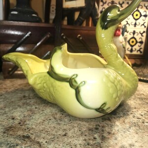 May include: A ceramic swan-shaped planter in shades of pale yellow and green. The swan has a long, curved neck and a wide, open basin for planting. Dark green vine-like details accent the body. The planter is likely vintage.