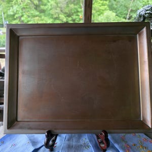 Puede incluir: Una bandeja rectangular de color bronce con un borde elevado y dos asas metálicas. La bandeja descansa sobre una tela azul con estampado floral. La bandeja probablemente se utiliza para servir alimentos o bebidas.