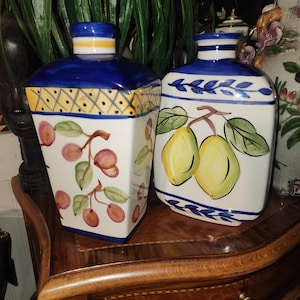 Può includere: Due vasi in ceramica con disegni dipinti in blu e bianco. Un vaso presenta un motivo geometrico giallo e blu con ciliegie rosse e rosa. L'altro vaso ha un motivo floreale blu e bianco con limoni gialli.