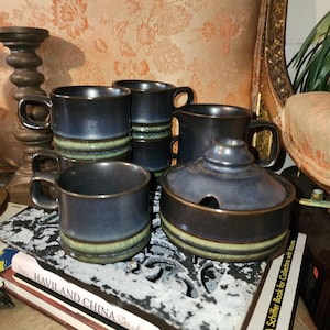 May include: A set of four blue and green ceramic mugs and a covered sugar bowl. The mugs have a dark blue glaze with a green band near the base. The sugar bowl has a matching design and a small spoon opening.