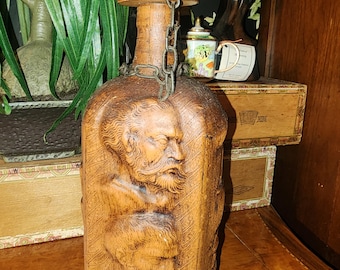 Könnte beinhalten: Eine Holz-Likörflasche mit einem geschnitzten Gesicht und einer Metallkette, die am Deckel befestigt ist. Die Flasche steht auf einer Holzoberfläche.