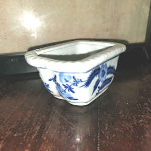 May include: A small white ceramic planter with a blue floral design. The planter has a rectangular shape with rounded edges.