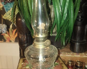 Puede incluir: Una lámpara de aceite de vidrio transparente con una base y una parte superior de metal color latón. La lámpara tiene un patrón floral decorativo en el depósito de vidrio. La lámpara está sobre una caja decorativa con un diseño floral.