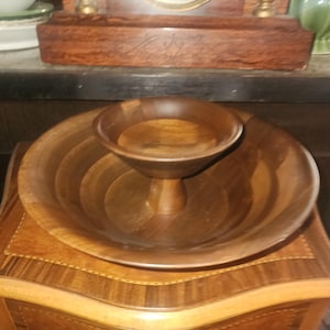 May include: A two-tiered wooden serving bowl with a dark brown finish. The bowl has a large, shallow base and a smaller, elevated bowl in the center. The wood grain is visible, adding to the rustic aesthetic. The bowl sits on a wooden cabinet.