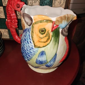 May include: A ceramic pitcher with a seahorse-shaped handle and fish-themed artwork. The pitcher features a red seahorse handle, and the body is decorated with colorful fish designs in shades of blue, orange, and green. The pitcher is on a red surface.