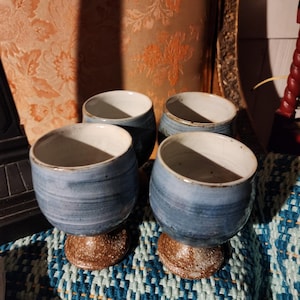 Puede incluir: Cuatro copas de cerámica hechas a mano. Cada copa presenta un cuenco esmaltado azul y blanco sobre una base marrón texturizada. Las copas están dispuestas sobre un textil tejido azul y blanco, con un fondo estampado.