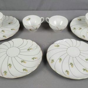 Könnte beinhalten: Ein Teeservice aus Porzellan bestehend aus zwei Teetassen und vier passenden Untertassen. Jedes Teil ist weiß mit einem Wirbelmuster aus dünnen Goldlinien und kleinen gelben Blumenmotiven. Die Untertassen haben gewellte Ränder und eine Vertiefung.