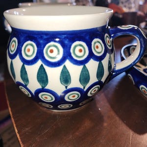 May include: A ceramic mug with a white interior and a blue exterior decorated with a repeating pattern of blue circles, green leaves, and orange dots. The handle is also blue with a matching design. The mug sits on a brown surface.