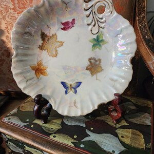 May include: A white, scalloped-edge decorative plate with iridescent sheen. The plate features hand-painted autumn leaves and a blue butterfly. A gold-colored handle with a hole is on the side. The plate is displayed on a decorative wooden stand.