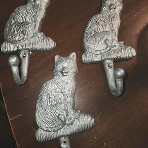 Puede incluir: Tres perchas de pared plateadas con forma de gato. Cada gancho presenta una figura detallada de gato sentado sobre una base decorativa, con un gancho curvo debajo. Los ganchos son de metal y están diseñados para colgar objetos.