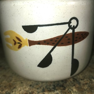 May include: White ceramic canister with a brown, yellow and black design of kitchen utensils. The lid is closed.