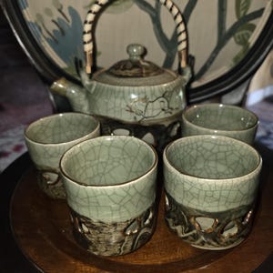 May include: A ceramic tea set featuring a teapot with a woven handle and four matching cups. The pieces have a light green crackle glaze on the upper portion and a dark, textured base with cutout details. The teapot includes a matching lid.