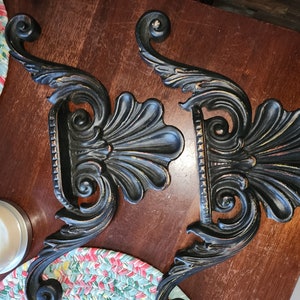 May include: Two black ornate metal wall brackets with a decorative scroll design. The brackets have a distressed finish and are perfect for adding a touch of elegance to any room.