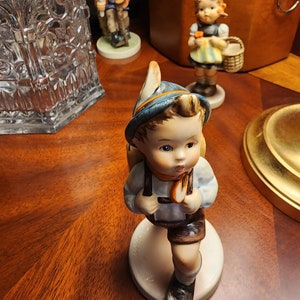 May include: A porcelain figurine of a young boy wearing a blue and white hat and carrying a backpack. He is wearing a blue and white shirt, brown shorts, and brown shoes. The figurine is standing on a white base.