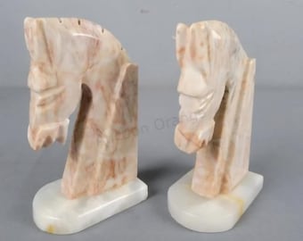 May include: Two marble horse head sculptures. The sculptures are in a light beige color with brown veining. Each horse head is mounted on a rectangular base. The sculptures are likely bookends or decorative items.