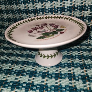 Puede incluir: Soporte para pasteles de cerámica blanca con una ilustración botánica de flores moradas y hojas verdes. El borde y la base están decorados con un patrón de hojas verdes. El soporte está sobre una superficie tejida turquesa y blanca.