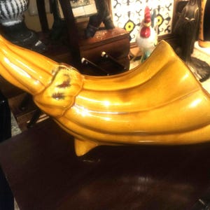 May include: A ceramic cornucopia in shades of yellow and brown. The horn-shaped vessel has a glossy finish and is decorated with ribbed detailing. The tip of the horn has a red accent. The cornucopia is placed on a dark brown surface.