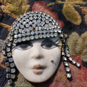 Könnte beinhalten: Vintage Brosche mit stilisiertem Gesicht, verziert mit einem Strass-Kopfschmuck und hängenden Strass-Akzenten. Das Gesicht ist mit zarten Merkmalen bemalt. Die Brosche befindet sich vor einem floral gemusterten Hintergrund.