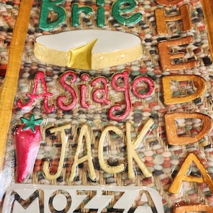 May include: A close-up of a decorative cheese board with the names of various cheeses in colorful, raised lettering. The cheeses include Brie, Asiago, Jack, and Mozzarella. A red pepper graphic is also present.