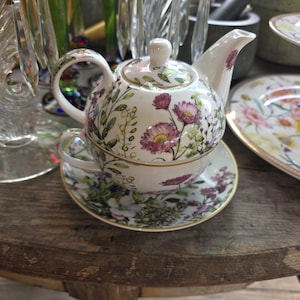 May include: A white porcelain tea set with a floral design featuring pink and purple flowers, green leaves, and gold trim. The set includes a teapot, teacup, and saucer, all stacked together on a wooden table.