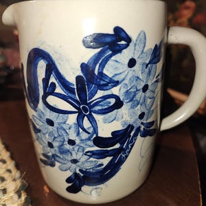 May include: White ceramic pitcher with a spout and handle, adorned with hand-painted blue floral and ribbon designs. The pitcher has a signature in cursive script. A charming piece for serving beverages.