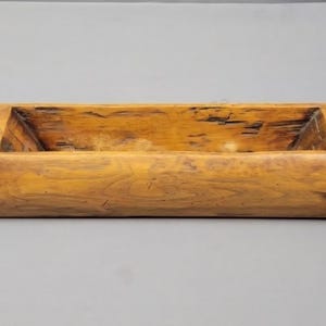 Könnte beinhalten: Eine rechteckige Teigschale aus Holz mit einer natürlichen hellbraunen Holzmaserung und einer hohlen Mitte. Die Schale verfügt über zwei glatte, abgerundete, dunkelbraune Holzgriffe, die an jeder Seite herausragen und eine lange Form bilden.