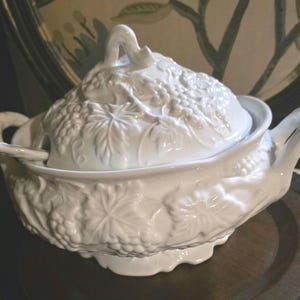 May include: A white ceramic tureen with a matching lid and ladle, featuring an embossed relief pattern of grape clusters and vine leaves. The tureen has two side handles and a scalloped base, with a decorative handle on the lid.