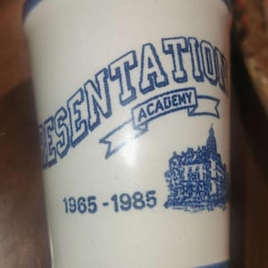 May include: White ceramic shot glass with blue trim and lettering. The text "PRESENTATION ACADEMY" arches across the front, with "ACADEMY" on a banner. Below is a building illustration and the dates "1965-1985".