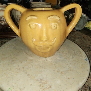 Könnte beinhalten: Eine gelbe Keramikkanne mit einem auf der Vorderseite modellierten Gesicht. Die Kanne hat zwei Henkel und eine breite Öffnung oben.